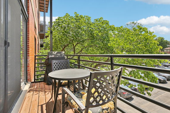 View from a balcony overlooking a parking area and trees, with outdoor furniture and a barbecue grill visible.