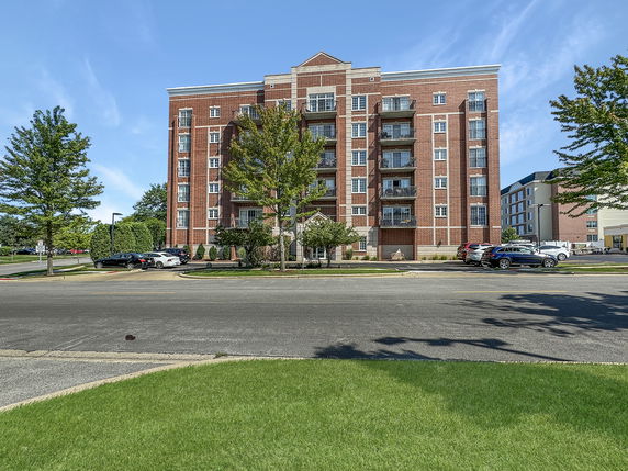 Front view of a multi-story brick building with balconies and a parking area in the foreground.