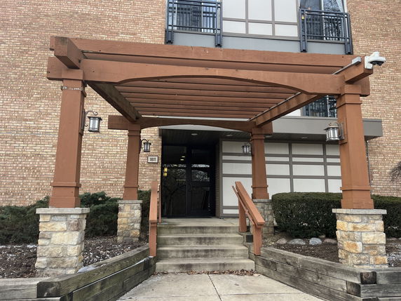 Front view of a building entrance with a wooden pergola and stone pillars.
