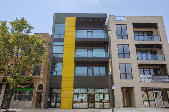 Front view of a modern multi-story building with glass balconies and a yellow panel.