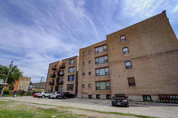 Front view of a mid-rise brick apartment building with balconies and parking area.