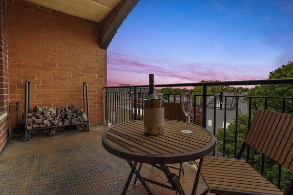 View from a balcony with a small table, chairs, and a railing overlooking rooftops and trees.