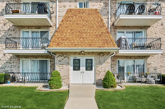Front view of a multi-story residential building with balconies and a distinctive sloped roof entrance.