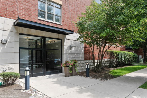 Front view of a brick building entrance with glass doors and surrounding greenery.