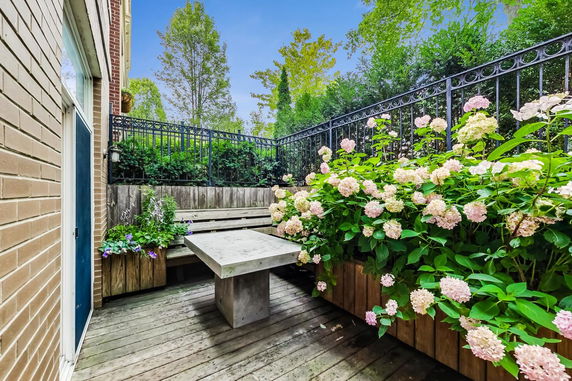View of a small outdoor patio area with wooden decking and a concrete table, surrounded by flowering plants and a metal fence.