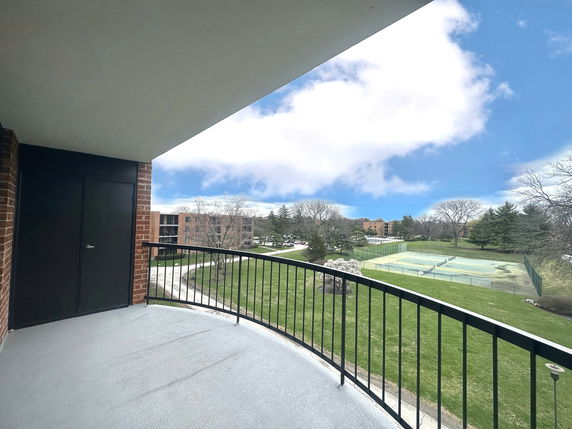 View from a covered balcony overlooking tennis courts and grassy area with buildings in the background.