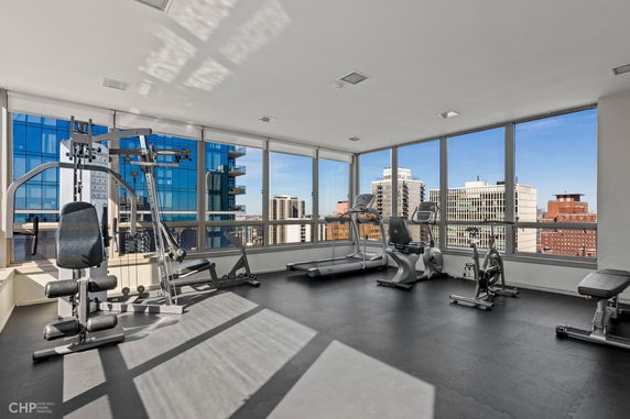 Interior of a gym with exercise equipment and large windows overlooking city buildings.