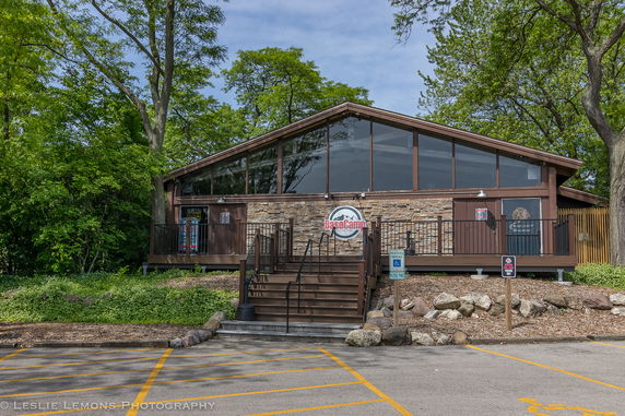 Front view of a building with large windows and a brown exterior, featuring stairs leading to the entrance. The building is surrounded by trees.
