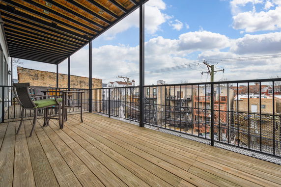 Wooden rooftop balcony with a table and chairs, overlooking adjacent buildings.