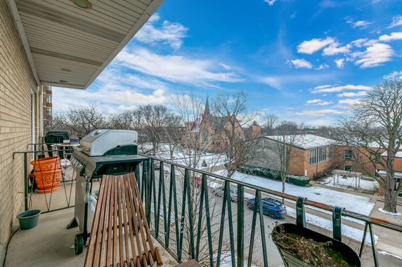 View from a balcony with a barbecue grill overlooking a neighborhood with trees and buildings in a snowy setting.