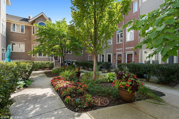 Front view of a row of townhouses with brick and siding exterior, featuring a landscaped garden and trees in the foreground.