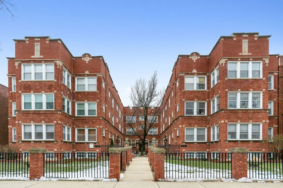 Front view of a multi-story brick apartment building with a central pathway and fenced entrance.