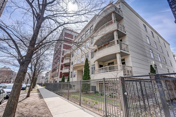 Front view of a multi-story apartment building with balconies and a fenced sidewalk.