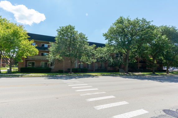 Front view of a multi-story apartment building with trees and a crosswalk in front.