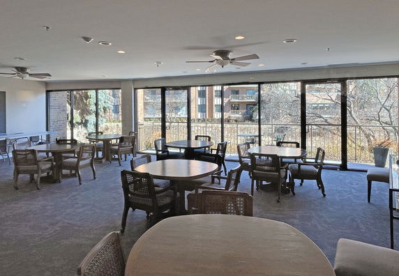 Indoor room with multiple round tables and chairs, large windows with a view outside.