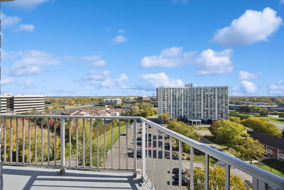 Panoramic view from a building balcony showing nearby high-rise buildings and a parking area.
