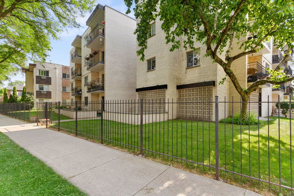 Front view of a multi-story apartment building with balconies and a fenced lawn.