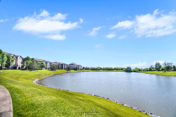 Wide view of a landscape featuring a lake surrounded by grass and buildings in the distance.