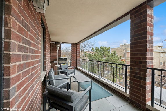 Covered balcony with seating and a grill, overlooking a residential area.