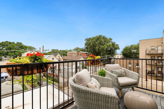 Outdoor seating area with chairs and plants on a balcony overlooking surrounding buildings and trees.