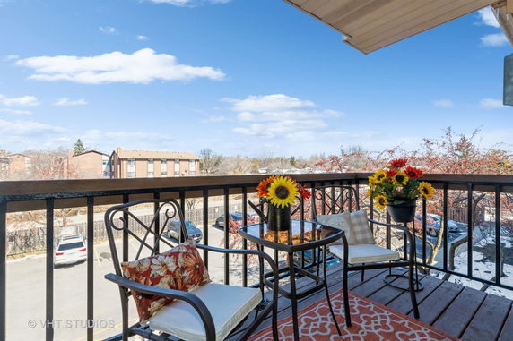 Balcony view showing outdoor seating with a table and potted flowers, overlooking a parking area and residential buildings.