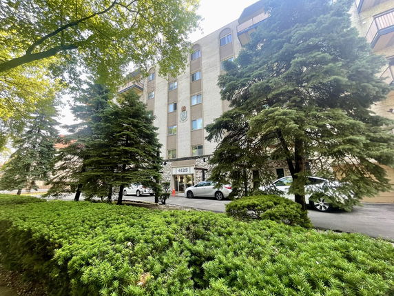 Front view of a multi-story brick building with balconies and a parking area in front.
