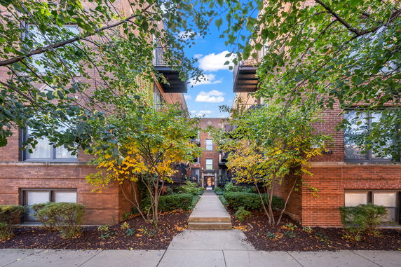 Front view of a multi-story brick apartment building entrance with trees and shrubs.
