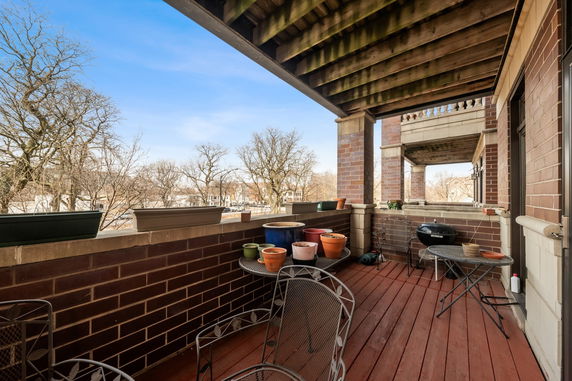 View from a covered balcony with potted plants and outdoor furniture overlooking trees and distant houses.