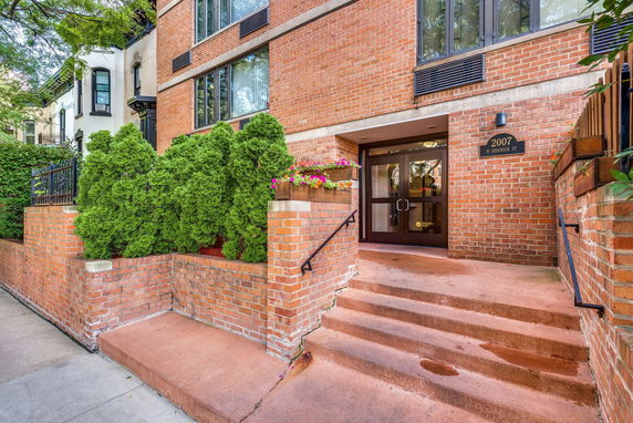 Front view of a brick building entrance with steps and surrounding greenery.