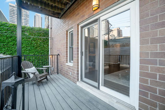 Outdoor patio with wooden deck and chairs, adjacent to a brick wall and overlooking surrounding buildings.
