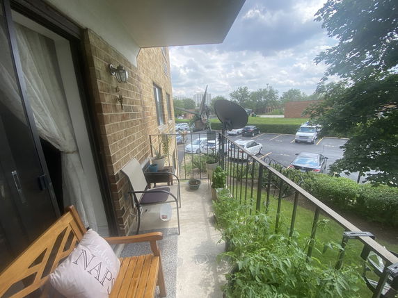 View from a balcony with chairs and plants, showing a parking area and nearby buildings.