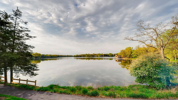 A scenic view of a calm lake surrounded by trees, with a paved path in the foreground.