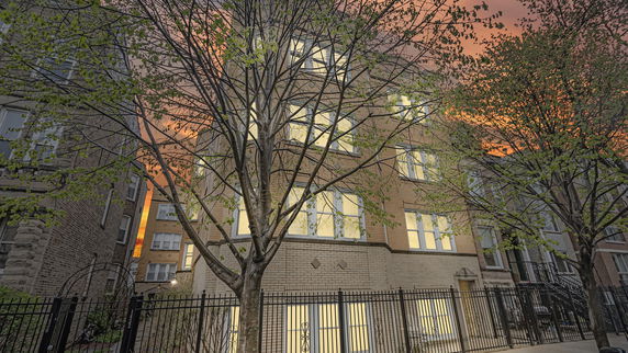Front view of a multi-story brick building with illuminated windows and a metal fence in the foreground.