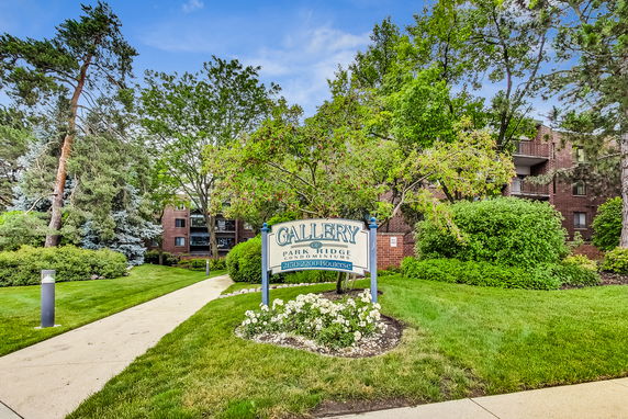 Front view of a brick condominium building with a walk path and sign reading 'Gallery Park Ridge Condominiums'.