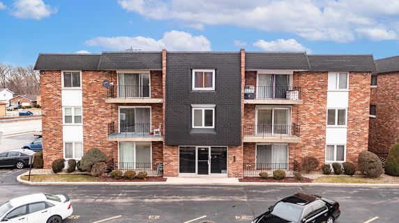 Front view of a three-story brick apartment building with balconies and a parking area.