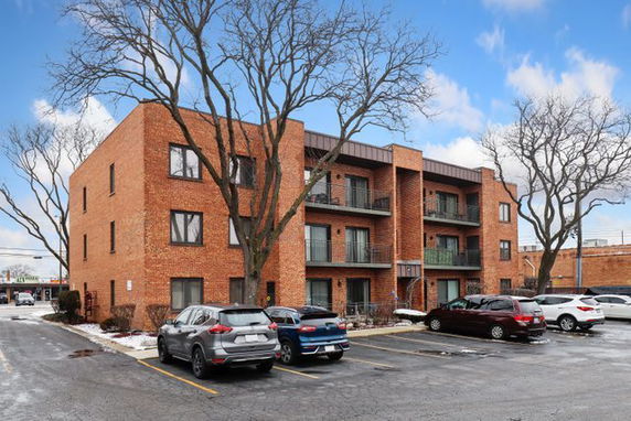 Front view of a three-story brick apartment building with balconies and a parking lot in front.