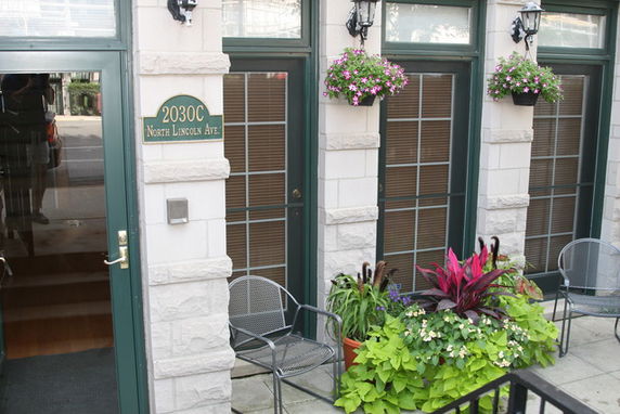 Front view of a building with large windows, flowers, and a seating area.