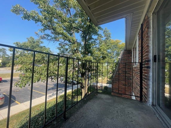 A view from a balcony with a railing, showing a parking area and trees nearby, with part of a brick building visible.