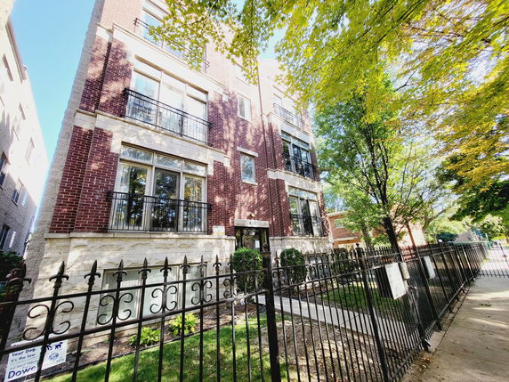 Front view of a multi-story brick building with large windows and a black iron fence.
