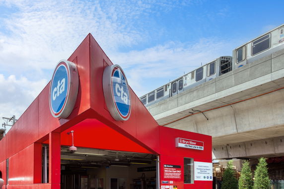Street-level view of an entrance to a public transport station with a train passing on an elevated track.