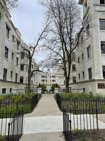 View of a multi-story building with a courtyard entrance surrounded by gardens and trees.