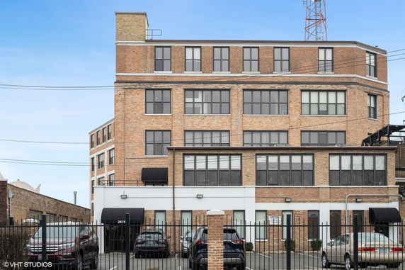 Front view of a multi-story brick building with large windows and a parking area in front.
