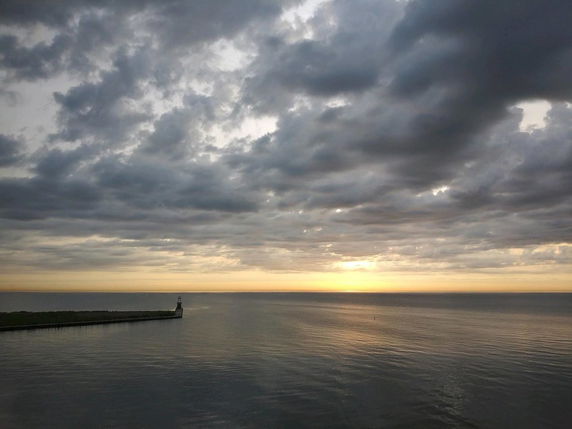 Wide view of ocean at sunset with cloudy skies and a distant pier.