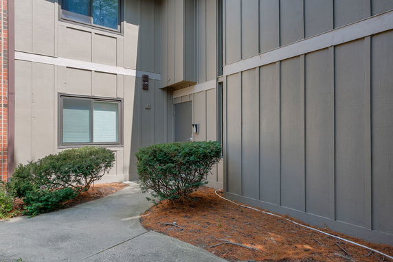 Exterior view of a building corner with two windows and shrubs along a pathway.