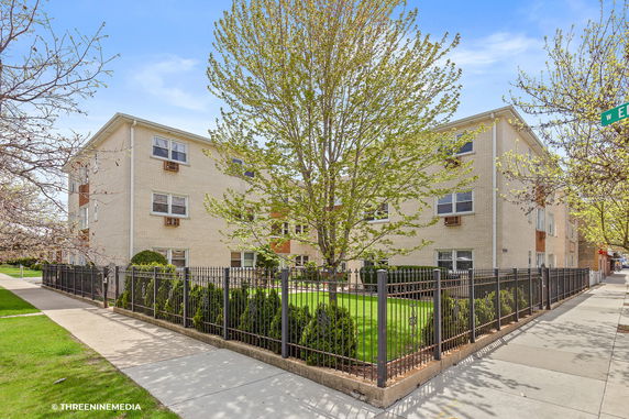 Front view of a three-story apartment building with light-colored brick exterior and fenced yard.