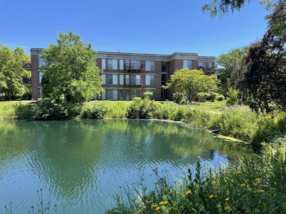 Front view of a three-story building with large windows and balconies, surrounded by greenery and a pond.