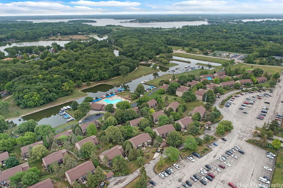 Wide aerial view of a residential area with houses, swimming pool, parking lots, and surrounding green landscape.
