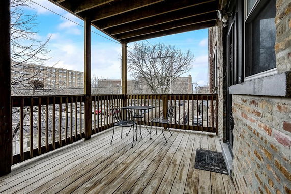 Wooden balcony with metal table and chairs, view of buildings and trees.
