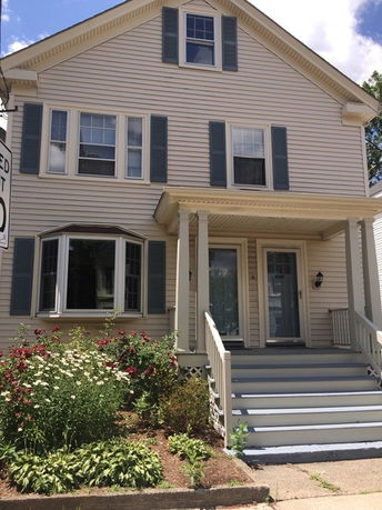 Front view of a two-story house with siding and shutters, featuring a covered porch entrance with steps.