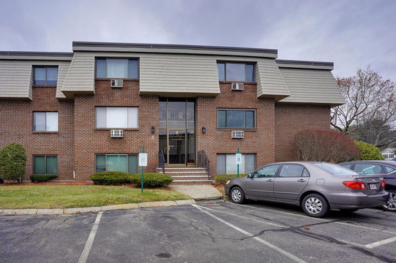 Front view of a multi-story brick building with parking spaces and windows with air conditioning units.